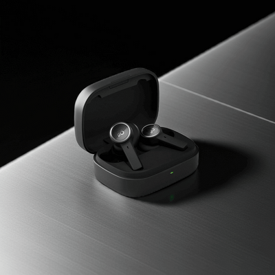 Nahaufnahme des geöffneten, mattschwarzen Ladeetuis der Bang & Olufsen BeoPlay EX Kopfhörer, platziert auf einer strukturierten, metallischen Oberfläche. Die beiden anthrazitfarbenen Ohrhörer mit ihren silbernen 'B&O'-Logoplaketten liegen im Etui, das eine kleine grüne Ladeanzeige zeigt